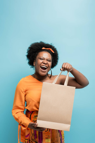 amazed african american body positive woman in orange dress holding shopping bag isolated on blue - Photo, Image
