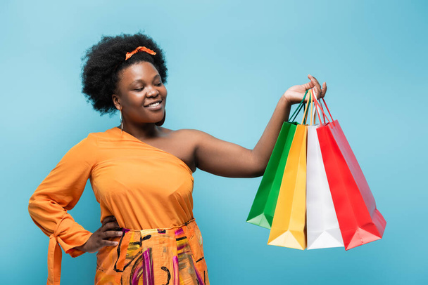 happy african american body positive woman holding shopping bags and posing with hand on hip isolated on blue - Fotoğraf, Görsel