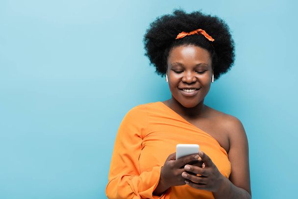 happy african american body positive woman in wireless earphones using smartphone isolated on blue - Foto, Imagen