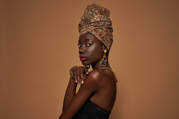Side view of fashionable black girl wearing traditional african outfit and accessories looking at camera. Pretty young slim woman. Isolated on orange background. Studio shoot. Copy space - Фото, изображение