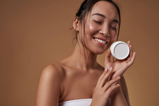Smiling elegant asian girl with closed eyes holding container with cosmetic cream. Beautiful young brunette slim woman wearing tank top. Body and skin care. Orange background. Studio shoot. Copy space - Φωτογραφία, εικόνα