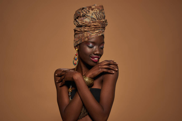 Smiling fashionable black girl wear african outfit and accessories hug herself. Young woman wearing tank top, turban, necklace, earrings and bracelet. Orange background. Studio shoot. Copy space - Foto, afbeelding