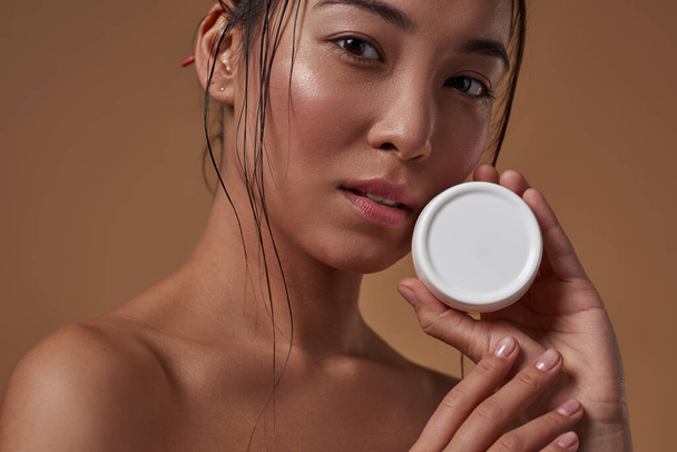 Partial of elegant asian girl holding container with cosmetic cream and looking at camera. Beautiful young brunette slim woman. Body and skin care. Isolated on orange background. Studio shoot - 写真・画像