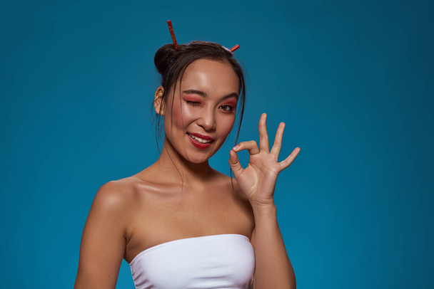 Fashionable asian girl winking, showing Ok sign and looking at camera. Pretty young brunette slim woman wearing tank top. Female beauty. Isolated on blue background. Studio shoot. Copy space - Photo, Image
