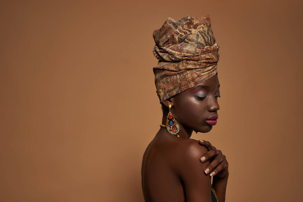 Side view of thoughtful black girl wearing traditional african outfit and accessories. Attractive young woman wear turban, necklace, earrings and bracelet. Orange background. Studio shoot. Copy space - Фото, зображення