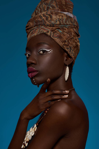 Cropped side view of fashionable black girl wearing traditional african outfit and accessories. Pretty young slim woman looking at camera. Female beauty. Isolated on blue background. Studio shoot - Foto, Imagem