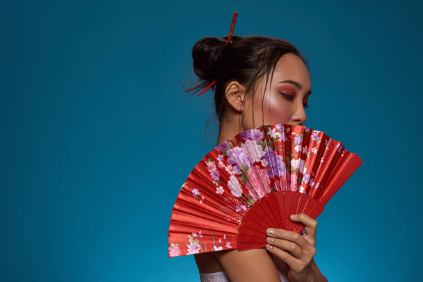 Side view of fashionable asian girl covering part of face with traditional handheld fan. Pretty young brunette slim woman wearing tank top. Female beauty. Blue background. Studio shoot. Copy space - Photo, Image