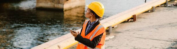 Black woman wearing helmet and vest using mobile phone while working in port - Foto, afbeelding