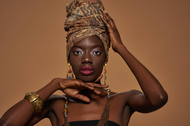Confident fashionable black girl wearing traditional african outfit and accessories posing and looking at camera. Pretty young slim woman. Isolated on orange background. Studio shoot. Copy space - Φωτογραφία, εικόνα