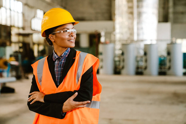 Black woman wearing helmet and vest smiling while working at factory - Fotó, kép