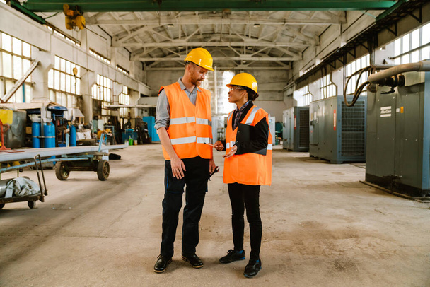 Multiracial man and woman in protective clothing talking while working at factory - Foto, Bild