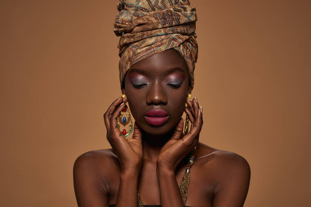 Portrait of elegant black girl wearing traditional african outfit and accessories. Beautiful young slim woman wearing turban, necklace and earrings. Isolated on orange background in studio. Copy space - Φωτογραφία, εικόνα