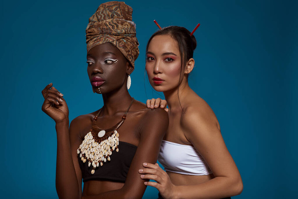 Multiracial girl friends in their national traditional styles. Black girl wearing african outfit and accessories looking away. Asian woman looking at camera. Blue background. Studio shoot. Copy space - 写真・画像