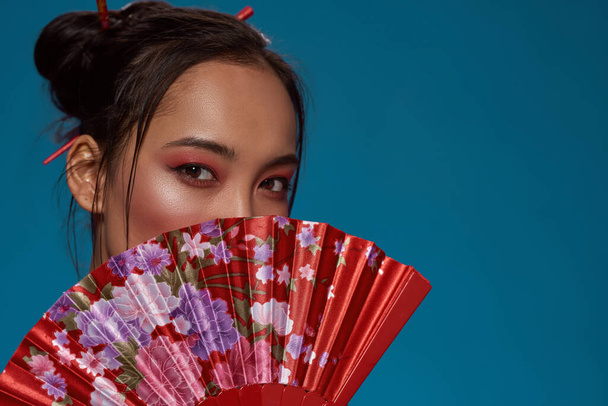 Partial of stylish asian girl covering half face with traditional handheld fan. Attractive young brunette woman looking at camera. Female beauty. Isolated on blue background. Studio shoot. Copy space - Foto, immagini