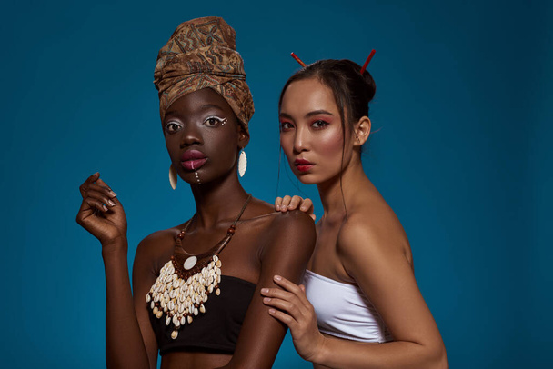 Multiracial girl friends in their national traditional styles looking at camera. Black girl wearing african outfit and accessories with asian woman. Isolated on blue background in studio. Copy space - Foto, immagini
