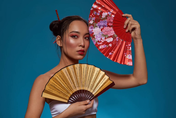 Serious stylish asian girl with traditional handheld fans looking at camera. Attractive young brunette slim woman wearing tank top. Female beauty. Isolated on blue background. Studio shoot. Copy space - Photo, Image