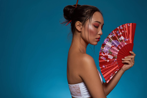 Side view of thoughtful stylish asian girl with traditional handheld fan. Attractive young brunette slim woman wearing tank top. Female beauty. Isolated on blue background. Studio shoot. Copy space - Photo, Image