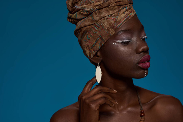 Cropped of stylish black girl. Attractive young slim woman wearing wearing traditional national african turban and earrings. Female beauty. Isolated on blue background. Studio shoot. Copy space - Foto, immagini