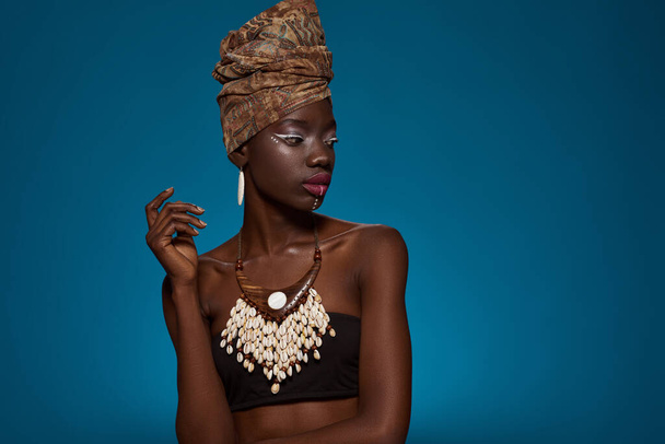 Focused fashionable black girl wearing african outfit and accessories. Pretty young slim woman wearing tank top, turban, necklace and earrings. Female beauty. Blue background. Studio shoot. Copy space - Photo, Image