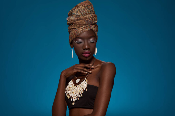 Thoughtful elegant black girl wearing african outfit and accessories. Beautiful young woman wearing tank top, turban, necklace and earrings. Female beauty. Blue background. Studio shoot. Copy space - Foto, Imagen