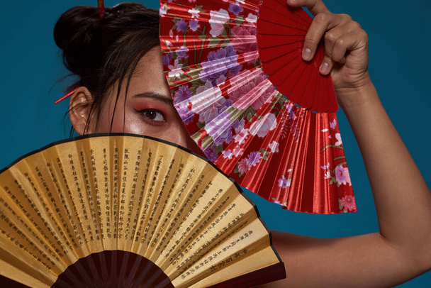 Elegant asian girl covering face with two traditional handheld fans. Beautiful young brunette slim woman looking at camera. Female beauty. Isolated on blue background. Studio shoot - Фото, зображення