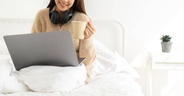 Jong glimlachend gelukkig mooi aziatisch vrouw ontspannen met behulp van laptop computer in de slaapkamer thuis. Jong creatief meisje werken en typen op toetsenbord.work vanuit huis concept - Foto, afbeelding