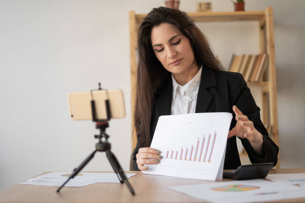 Positive businesswoman recording video content for her blog at office, streaming online podcast on mobile phone on tripod shows and talks about savings in budget. Technology and blogging concept. - Fotoğraf, Görsel