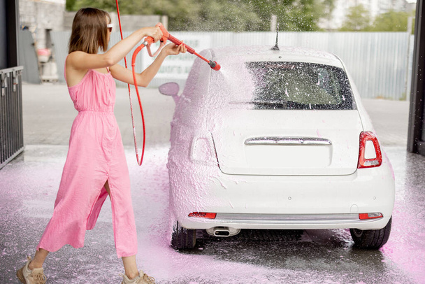 Young woman in pink dress washing her tiny car with nano foam at car wash during summer time. Easy and beautiful self-service at car wash concept - Photo, Image