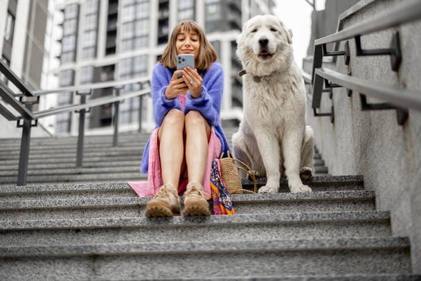 Vrolijke vrouw zit met haar schattige hond op de trap en het gebruik van de telefoon in de moderne woonwijk. Vrije tijd en vriendschap met huisdieren concept - Foto, afbeelding