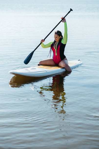 Young Woman with Dreadlocks in Swimwear Sitting on the Sup Board, Female Paddleboarding on Lake at Sunrise - Fotó, kép