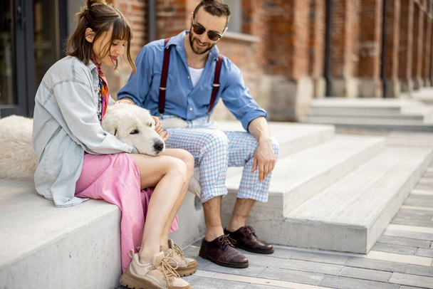 Young stylish couple sit together and have fun with their white dog on a street. Young hipsters hang out together near office outdoors - Foto, imagen