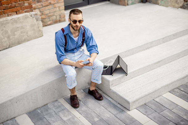 El hombre con estilo utiliza el teléfono inteligente y cargarlo desde el panel solar portátil mientras está sentado en la calle al aire libre. Moderno estilo de vida sostenible y energías renovables - Foto, imagen