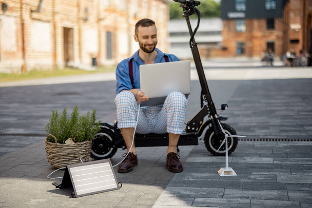 Stylish man works on laptop computer and charge it with solar panels while sitting on electric scooter at office district. Concept of sustainable lifestyle and green renewable energy - Zdjęcie, obraz