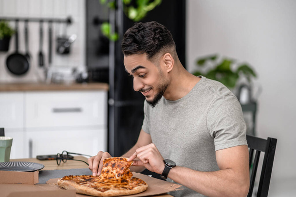 Ест дома. Счастливый молодой бородатый мужчина дотрагивается до пиццы, сидя боком перед камерой в квартире - Фото, изображение