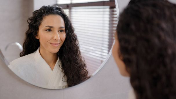 Close-up serious pensive young hispanic woman looking in mirror in bathroom contemplating future cute curly brunette girl in bathrobe admires reflection natural beauty daily morning routine concept - Foto, Bild