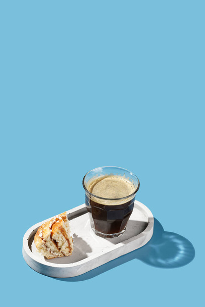 Hard light photo of a glass with espresso and a piece of french cinnamon pastry on an oval concrete tray on blue background - Photo, Image