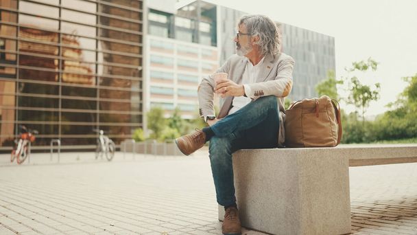 Mature businessman with beard in eyeglasses wearing gray jacket holding takeaway coffee. Middle aged manager enjoying morning coffee while sitting outside the office - Foto, Imagen