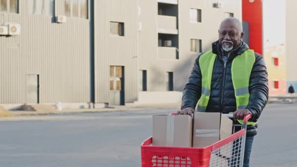 Mature african american deliveryman using handcart with cardboard boxes professional courier loader in uniform transports trolley with parcels to car deliver purchases efficiently delivery service - Materiał filmowy, wideo