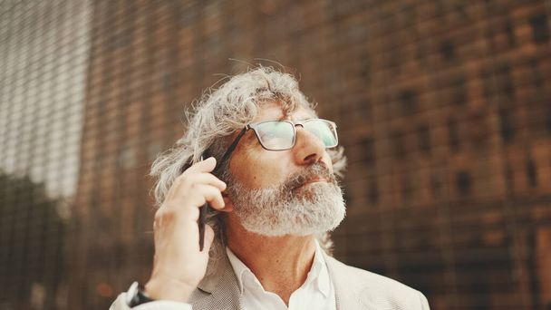 Clouse up, mature businessman with beard in eyeglasses wearing gray jacket is using cell phone. Middle aged manager talking on smartphone while sitting outside office - 写真・画像