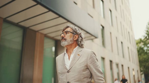 Mature businessman with beard in eyeglasses wearing gray jacket walks down the street past modern buildings - Photo, image