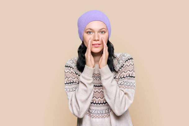 Young attractive girl in winter sweater hat telling secret rumors, on light brown color background. Cute teenage female looking at camera, whispering holding her hands near her lips - Fotó, kép