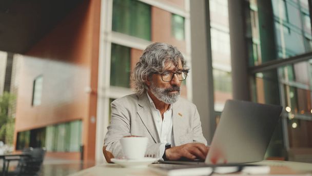 Mature businessman with beard in eyeglasses wearing gray jacket sits on cafe. Middle aged manager successful man user on laptop pc computer sit at cafe outdoors - Photo, image