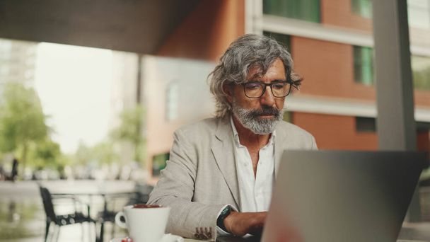 Clouse up, mature businessman with beard in eyeglasses wearing gray jacket sits on cafe. Middle aged manager successful man user on laptop pc computer sit at cafe outdoors - Photo, Image