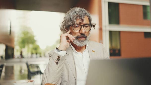 Clouse up, mature businessman with beard in eyeglasses wearing gray jacket sits on cafe. Middle aged manager successful man user on laptop pc computer sit at cafe outdoors - Foto, imagen