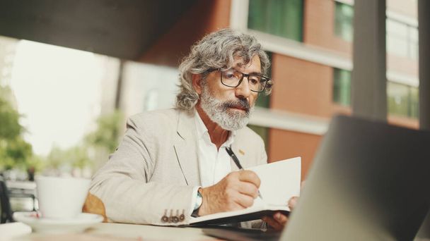 Mature businessman with beard in eyeglasses, user on laptop computer and taking notes, sitting in outdoor cafe. Successful man sitting at table in cafe in nature Mobile office freelancer - Φωτογραφία, εικόνα