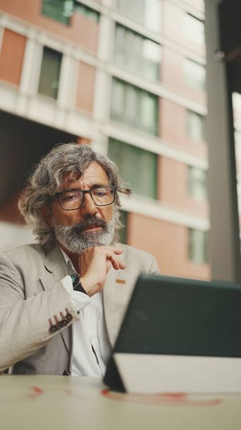 Mature businessman with beard in eyeglasses wearing gray jacket, working on tablet, sitting in an outdoor cafe. Successful man sitting at table in cafe in nature Mobile office freelancer - 写真・画像