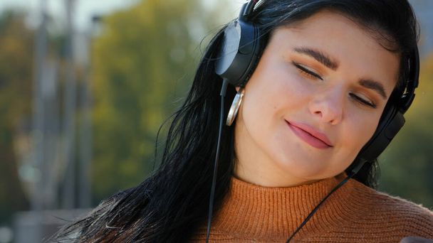 Portrait young happy woman standing outdoors in headphones listening favorite music using smartphone carefree hispanic girl smiling dancing to pleasant song melody audio sound female enjoying life - Photo, Image