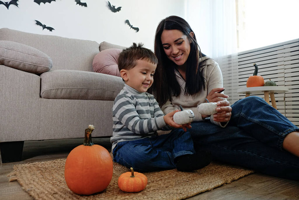 Young beautiful woman with her three year old son preparing decorations for halloween at home. Mom spending quality time with her child, messing around with pumpkins. Close up, copy space, background. - Photo, image