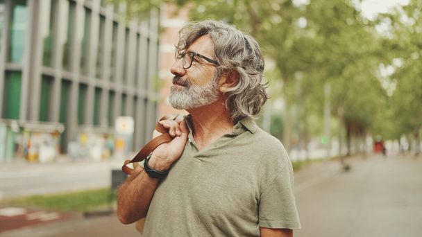 Middle-aged man with gray hair and beard walks and looks around. Mature gentleman in eyeglasses with bag on his shoulder walks through the square on the cityscape background - Фото, изображение
