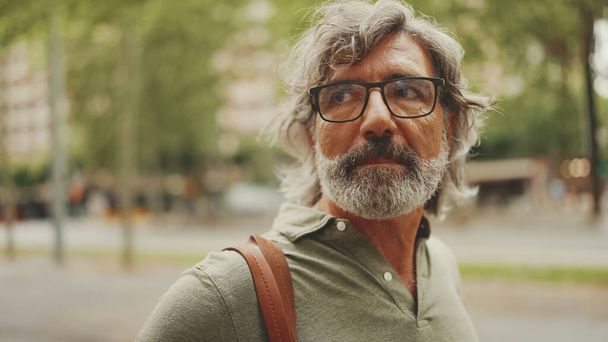 Middle-aged man with gray hair and beard walks and looks around. Mature gentleman in eyeglasses with bag on his shoulder walks through the square on the cityscape background - Fotografie, Obrázek